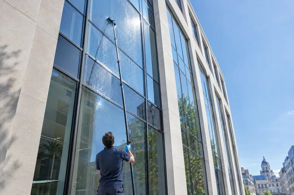 Un laveur de vitres nettoie depuis le sol les hautes fenêtres d’un immeuble de bureaux avec une perche à eau pure
